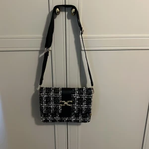 Svart och vit rutig axelväska från Lycke Oslo - Säljer en snygg axelväska från Lycke Oslo i svart och vitt rutigt tyg. Väskan har guldfärgade detaljer och justerbar axelrem. Perfekt för att lyfta din outfit med en trendig accessoar.