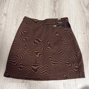 Brun mönstrad kjol från H&M Divided - Säljer en brun kjol med svart vågigt mönster från H&M Divided. Kjolen är kort och har en snygg detalj med utskärning och en silver ring vid midjan