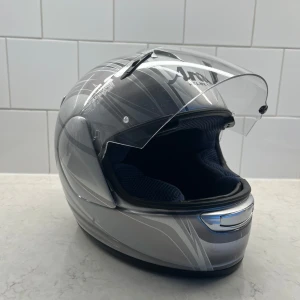 Arai Chaser MC-hjälm i silver och grå - Snygg integralhjälm från Arai i modellen Chaser. Hjälmen är silverfärgad med gråa och vita grafiska linjer, har visir och vadderad insida för extra komfort. Perfekt för motorcykelåkning och har klassisk Arai-logga på flera ställen.        Orginal pris 9000kr