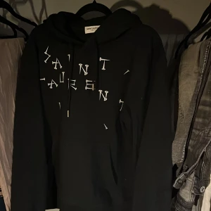 Svart hoodie från Saint Laurent med tryck - Säljer en svart hoodie från Saint Laurent med unikt vitt tryck framtill där bokstäverna är utspridda. Klassisk huva med dragsko och Saint Laurent-detalj på snöret. Perfekt för dig som gillar streetwear och exklusiva märken.