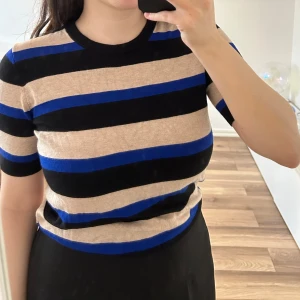 Randig topp från Zara - Säljer en kortärmad topp från Zara med breda ränder i blått, svart och beige. Toppen har rund halsringning och knappar bak i nacken. Mjukt material.