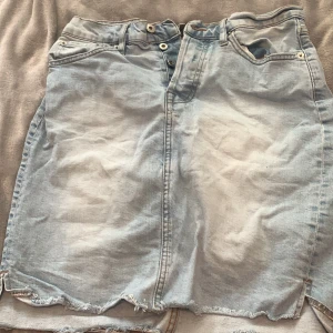 Ljusblå jeanskjol från 157 - Säljer en ljusblå jeanskjol från 157 med rå kant nertill och klassiska fickor både fram och bak. Kjolen har knappar som man knäpper där fram samt en rak passform. Perfekt för dig som gillar en avslappnad och trendig stil.