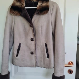 Beige mockajacka med pälsdetaljer - Snygg beige mockajacka med bruna pälsdetaljer på krage och ärmslut. Jackan har knappar framtill och två fickor med mörka detaljer. Perfekt för dig som vill ha en stilren och varm jacka med lyxig känsla.