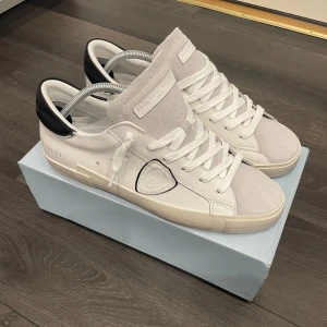 Vita sneakers från Philippe Model - Säljer ett par snygga vita Philippe Model sneakers med svarta detaljer på hälen och vit snörning. Skorna har en kombination av mocka och skinn, skorna är HELT NYA! och aldrig använda bara testade. Priset är ej hugget i sten så kom gärna med förslag! Säljer de då jag behöver pengar😞