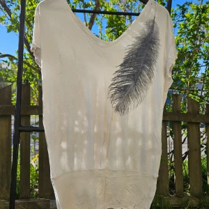 Vit t-shirt med fjädertryck - Vit t-shirt med djup v-ringning och ett stort svart fjädertryck på framsidan. T-shirten har korta ärmar och är tillverkad i ett mjukt material som känns skönt mot huden. Perfekt för en avslappnad stil.