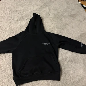 Svart hoodie från Essentials - Säljer en svart hoodie från Essentials med tryckt logga på bröstet och ärmen. Tröjan har huva och en stor magficka framtill. Perfekt för en avslappnad och stilren look.