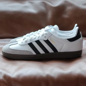 Adidas Samba sneakers i vitt och svart - Klassiska Adidas Samba sneakers med vit ovandel, svarta ränder och detaljer samt grå mocka vid tån. Skon har brun gummisula och snörning framtill. Perfekt för en sportig och stilren look. Köpta på junkyard för 1500kr