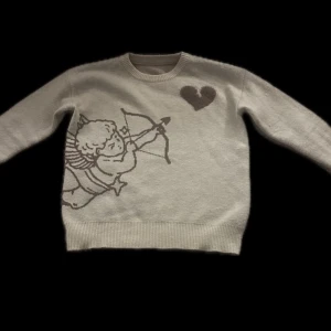 Vit stickad tröja med ängelmotiv och text - Vit stickad tröja med ett motiv av en ängel som skjuter pil och ett hjärta på framsidan. På baksidan står texten 'I WOULD RATHER NOT LOVE' och dekorativa stjärnor. Rund hals och ribbade muddar.