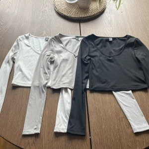 3-pack långärmade croppade toppar från SHEIN - Säljer tre långärmade croppade toppar från SHEIN i färgerna svart, grå och vit. Alla har rundad v-ringning och är tillverkade i ett ribbat, stretchigt material. Perfekta att matcha med jeans eller kjol för en snygg look.