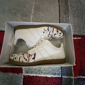 Gats - Säljer ett par vita sneakers med färgglatt camouflagemönster i blått, rött och beige på tån och sidorna. Skorna har vita snören och en brun sula. Perfekta för dig som vill sticka ut med din stil.