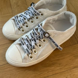 Vita sneakers från Christian Dior - Säljer ett par vita Walk’n’ Dior sneakers från Christian Dior med unika snören där det står 'CHRISTIAN DIOR J'ADIOR'. Skorna har en platt sula med svart stjärndetalj undertill och är tillverkade i canvasmaterial. Märkeslogga finns även på hälen.  