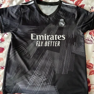 Real Madrid Benzema fotbollströja - Snygg svart Real Madrid fotbollströja med vitt tryck, klubbmärke på bröstet och 'Emirates Fly Better' på framsidan. Baksidan har 'Benzema' och nummer 9. Tröjan har korta ärmar och ett modernt grafiskt mönster.