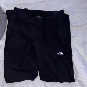 Svarta byxor från The North Face - Svarta byxor från The North Face med rakt snitt och diskret logga på låret. Byxorna har dragkedja och knapp framtill samt sidofickor. Perfekta för vardagsbruk eller utomhusaktiviteter.