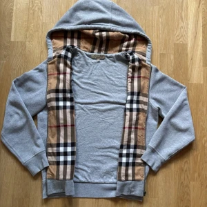 Hoodie från Burberry | ev byte - Säljer en grå hoodie från Burberry med dragkedja och ikoniskt beige, svart och röd rutigt foder i huvan och längs insidan. | ev byte