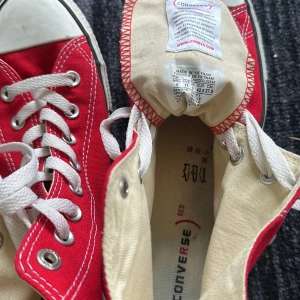 Röda Converse All Star high-top sneakers - M9621, Säljer ett par klassiska röda Converse All Star high-top sneakers med vita snören och gummitå. Skorna har vit sula med svart rand och den ikoniska Converse-loggan på sidan. Perfekta för dig som gillar en tidlös och cool stil.