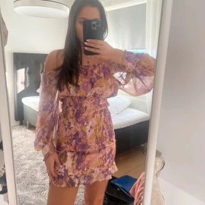 Blommig offshoulder klänning med volang - Säljer en superfin kort blommig klänning med offshoulder och långa, transparenta ärmar. Klänningen har volangdetaljer och smockad midja för en snygg passform. Perfekt för sommarens alla tillfällen!