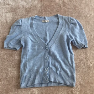 Ljusblå stickad kofta med puffärm - Säljer en ljusblå stickad kofta med korta puffärmar och v-ringning. Koftan har tre knappar framtill och bred ribbad kant nedtill och vid ärmslut. Aldrig använd. 💙