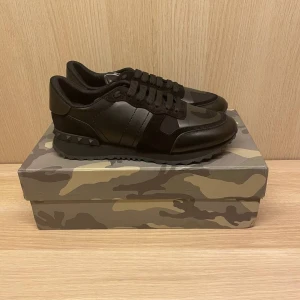 Svarta rockrunners sneakers från Valentino - Säljer ett par svarta Valentino sneakers med kamouflagemönster och detaljer i skinn och mocka. Skorna har snörning, rund tå och en platt sula med nitar på hälen. Perfekta för dig som gillar stilrena och exklusiva sneakers. Storlek 43. Priset ej skrivet i sten men var rimliga