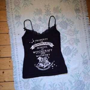 Svart Hogwarts linne med spets - Svart linne med tunna axelband och spetsdetaljer upptill. Tryck framtill med texten 'Property of Hogwarts School of Witchcraft and Wizardry' och Hogwarts-emblem. Perfekt för Harry Potter-fans!