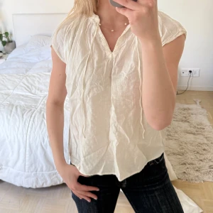 Ljusbeige kortärmad blus i luftig bomull - Säljer en ljusbeige blus med korta ärmar från Uniqlo. Tillverkad i tunn, luftig bomull – perfekt för varmare dagar. Blusen har en avslappnad passform, lätt rynkade detaljer vid halsringningen och ett knytband framtill. Enkel att styla med både jeans och kjol för en fräsch och ledig look.