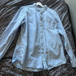 Ljusblå skjorta med knappkrage - Klassisk ljusblå skjorta med lång ärm och knappkrage ifrån dressmann. Skjortan har en bröstficka med knapp och stängs med vita knappar framtill. Perfekt för en clean och stilren look.