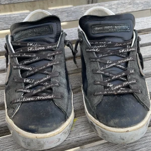 Svarta Philippe Model sneakers med vita detaljer - Säljer ett par svarta Philippe Model sneakers med vita detaljer och logga på sidan. Skorna har snörning med text på skosnörena och en platt sula. Perfekta för dig som gillar streetstyle och vill ha något unikt. 