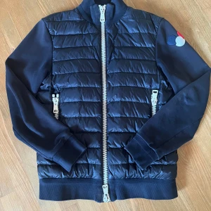 Moncler cardigan - En snygg marinblå Moncler cardigan i storlek S. Små tecken på blekning kan förekomma, samt två defekter, en på vardera arm (ett exempel visas på bilden). Samt har de vita framtiderna tappat lite färg. Snygg tripple badge som är ganska ovanlig 