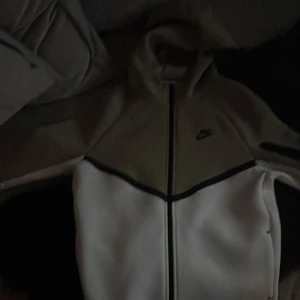 Nike hoodie i beige och vitt - Säljer en Nike hoodie i beige och vitt med dragkedja och huva. Jackan har lång ärm och svart Nike-logga på bröstet. Perfekt för dig som gillar sportig stil och vill ha något bekvämt och snyggt. Materialen på hoodin är gjort av fleece