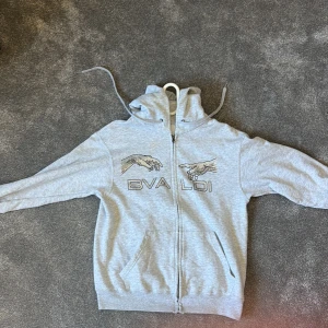 Ljusgrå hoodie med tryck från BVALDI - Säljer en ljusgrå hoodie med dragkedja från BVALDI. Tröjan har tryck med två händer och text på bröstet, huva med dragsko och två fickor framtill. Perfekt för en avslappnad stil.