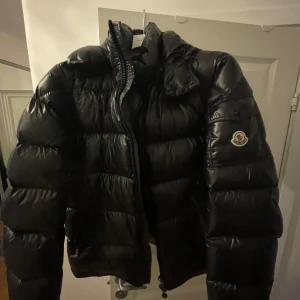 Svart dunjacka från Moncler - Säljer en svart dunjacka från Moncler med hög krage och huva. Jackan har lång ärm, dragkedja framtill och Moncler-logga på ärmen. Klassisk pufferstil som håller dig varm under kalla dagar.