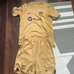 Gul FC Barcelona fotbollströja med shorts - Säljer ett gult FC Barcelona matchställ från Nike med kortärmad tröja och shorts. Tröjan har klubbmärke, sponsorlogga och regnbågsfärgade detaljer vid ärmslut. Nummer 4 och namnet R. Araujo på ryggen.