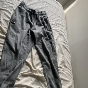 Grå mjukisbyxor från Under Armour Size S I Herr - Grå mjukisbyxor från Under Armour med elastisk midja och dragsko. Byxorna har en klassisk passform, sidofickor och en bakficka. Materialet är mjukt och skönt, perfekt för chill eller träning. Diskret logga på vänster ben.