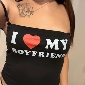 Svart tubtopp med text I ❤️ MY BOYFRIEND - Svart tubtopp med vita bokstäver och ett rött hjärta på bröstet där det står 'I ❤️ MY BOYFRIEND'. Axelbandslös modell med smala band på sidorna. Perfekt statement-plagg för dig som vill sticka ut.