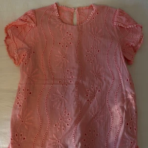 Rosa blus med broderade blommor - Supersöt rosa blus med korta ärmar och broderade blommor samt vågiga hålbroderier. Blusen har rund halsringning och knäppning bak i nacken. Perfekt för dig som gillar romantiska detaljer och en luftig känsla.