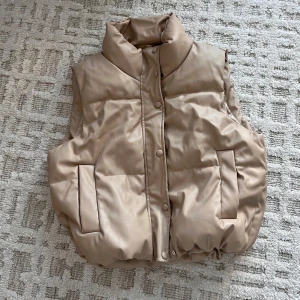 Beige dunväst med hög krage - Snygg beige dunväst med hög krage och två stora fickor framtill. Västen har både dragkedja och tryckknappar för extra stil och funktion. Den har en puffig, loose passform som ger en cool och avslappnad vibe. Perfekt att slänga över en hoodie eller tröja när det är kyligt ute.