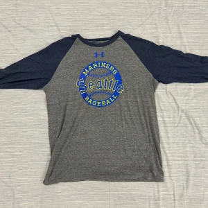 Seattle Mariners långärmad tröja XL - Snygg långärmad tröja från Under Armour i grått med marinblå ärmar och tryck med Seattle Mariners Baseball i blått och gult. Tröjan har loose fit och är gjord i mjuk bomullsmix, perfekt för sport eller chill. Klassisk rund hals och HeatGear-material.