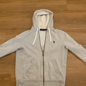 Grå hoodie från Polo Ralph Lauren - Snygg ljusgrå hoodie från Polo Ralph Lauren med dragkedja och huva med vita snören. Tröjan har det klassiska Polo-logot broderat på bröstet och två fickor framtill.