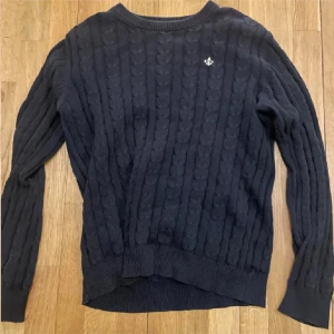 Mörkblå stickad tröja med emblem - Snygg mörkblå stickad tröja från Gant med klassiskt kabelstickat mönster och ett litet silverfärgat emblem på bröstet. Tröjan har rund hals och långa ärmar, perfekt för dig som gillar stilren och tidlös design.