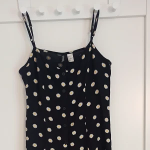 Svart blommig miniklänning från H&M Divided - Supersöt svart kortklänning från H&M Divided med vita och gula blommor🌼 Klänningen har knappar framtill och är tillverkad i mjuk viskos som känns skön mot huden. Perfekt för dig som gillar blommiga klänningar! 