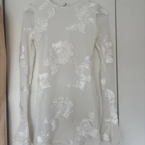 Vit kort klänning med blommig spets - Supersnygg vit kort klänning med långa ärmar med volang. Klänningen har stora blommiga spetsdetaljer och en öppen rygg. Passformen är slim och tyget känns lätt och festligt. Perfekt för flera olika tillfällen. Endast använd 1 gång 🤍