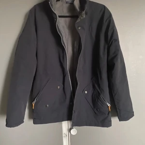 Svart vindjacka med fleecefoder - Svart vindjacka från Polarland med grått fleecefoder. Jackan har huva, dragkedja och tryckknappar framtill samt två stora fickor med dragkedja och knappstängning. Perfekt för kyliga dagar när du vill hålla dig varm och bekväm.