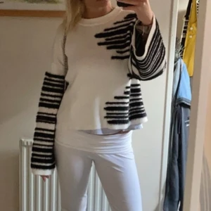 Vit och svart randig stickad tröja - Säljer en vit stickad tröja med svarta, oregelbundna ränder på ärmar och framsida. Tröjan har lång ärm, ribbad struktur och en loose fit. Perfekt för dig som gillar en unik och trendig look med grafiska detaljer.