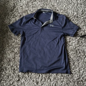 Marinblå pikétröja från Under Armour - Aldrig använd. Snygg marinblå pikétröja från Under Armour med klassisk krage och korta ärmar. Tröjan är tillverkad i ett lätt och ventilerande syntetmaterial, perfekt för träning eller chill dagar. Under Armour-logga på bröstet och grå detalj på insidan av kragen.