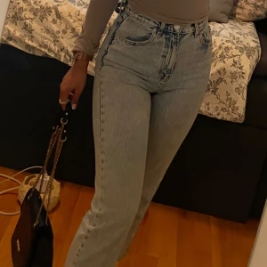 Ljusblå raka jeans med hög midja - Säljer ett par ljusblå jeans med hög midja och raka ben. Jeansen har klassisk femficksdesign, dragkedja och knapp framtill. De är tillverkade i jeansmaterial och har en snygg tvättad look som ger en cool vintagekänsla.