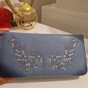 Blå plånbok med vingar och nitar - Snygg ljusblå plånbok med silvriga nitar som bildar ett coolt vingmotiv på framsidan. Plånboken har dragkedja runtom och flera fack för kort och mynt på insidan. Perfekt för dig som gillar detaljer och vill ha något unikt i väskan.