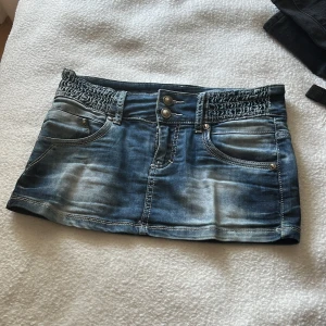 Blå jeanskjol från Toxik3 - Supersnygg kort jeanskjol från Toxik3 med cool tvättad blå färg och vita sömmar. Kjolen har två framfickor, två bakfickor och bred smock i midjan med dubbla knappar framtill. Perfekt för dig som gillar streetstyle och denim.