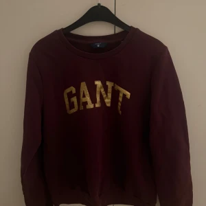 Vinröd sweatshirt från GANT i M - Snygg vinröd sweatshirt från GANT i storlek M (passar  om du har S) mermed guldfärgat tryck på bröstet. Tröjan har rund hals, långa ärmar och är gjord i mjuk bomull. Perfekt för dig som gillar klassisk stil med en touch av lyx.