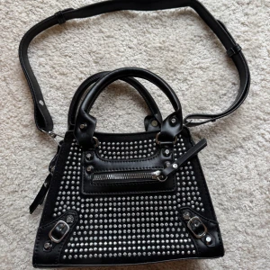 Svart axelväska med nitar och dragkedja - Cool svart axelväska i skinnimitation med silvriga nitar på framsidan och detaljer som spännen och dragkedja. Väskan har både handtag och justerbar axelrem, perfekt för att även bära crossbody. Stilren baksida och rymligt fack med dragkedja upptill.Ej använd. Mått 22X16x8 cm. 