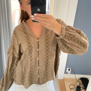 Khaki broderad blus från Zara - Säljer en  från Zara med v-ringning och knappar framtill. Blusen har broderade detaljer i vertikala rader och ballongärmar med knapp vid ärmslut. Endast prövad 