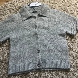 Grå stickad kofta från Gina Tricot - Kortärmad stickad kofta i ljusgrå melange från Gina Tricot. Modellen har klassisk krage, ribbade muddar och knappar framtill. Perfekt att slänga över en t-shirt för en chill look. Mjuk och skön känsla, passar dig som gillar enkel och clean stil.